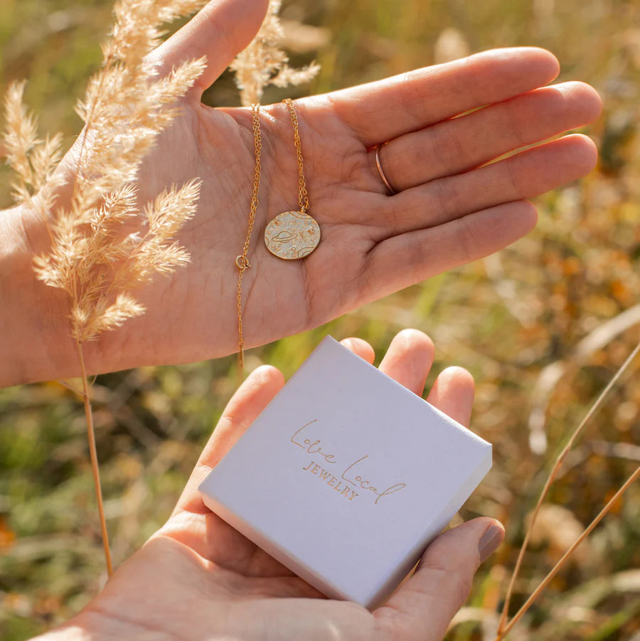 Collier doré motif planètes tenu dans la main avec coffret cadeau en arrière-plan naturel, idée cadeau femme.